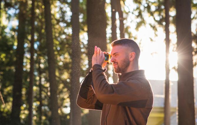 A male student wearing a brown jacket standing in a forested area while looking through a small yellow device that he's holding up to his eye