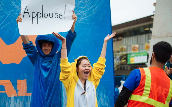 Two UBC students in animal onesies in front of Storm the Wall, with one student holding up a sign reading Applause.